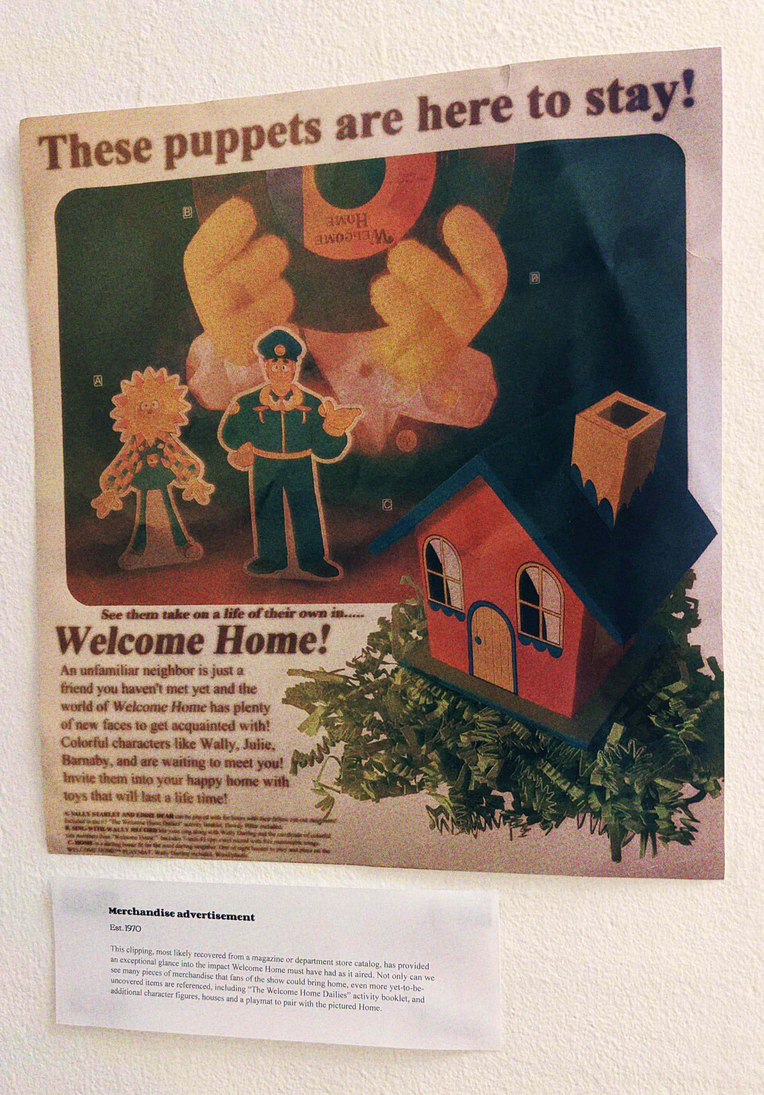 An advertisement hung on a wall, the ad shows Wally's puppet hands holding a vinyl record, Sally and Eddie cutouts, and a toy of Home. The ad reads: 'These puppets are here to stay! See them take on a life of their in... Welcome Home! An unfamiliar neighbor is just a friend you haven't met yet and the world of Welcome Home has plenty of new faces to get acquainted with! Colorful characters like Wally, Julie, Barnaby, and are waiting to meet you! Invite them into your happy home with toys that will last a life time!' A exhibit card under the ad reads: 'Merchandise advertisement. Est. 1970. This clipping, most likely recovered from a magazine or department store catalog, has provided an exceptional glance into the impact Welcome Home must have had as it aired. Not only can we see many pieces of merchandise that fans of the show could bring home, even more yet-to-be-uncovered items are refenced, including 'The Welcome Home Dailies' activity booklet, and additional character figures, houses, and a playmat to pair with the pictured Home.'