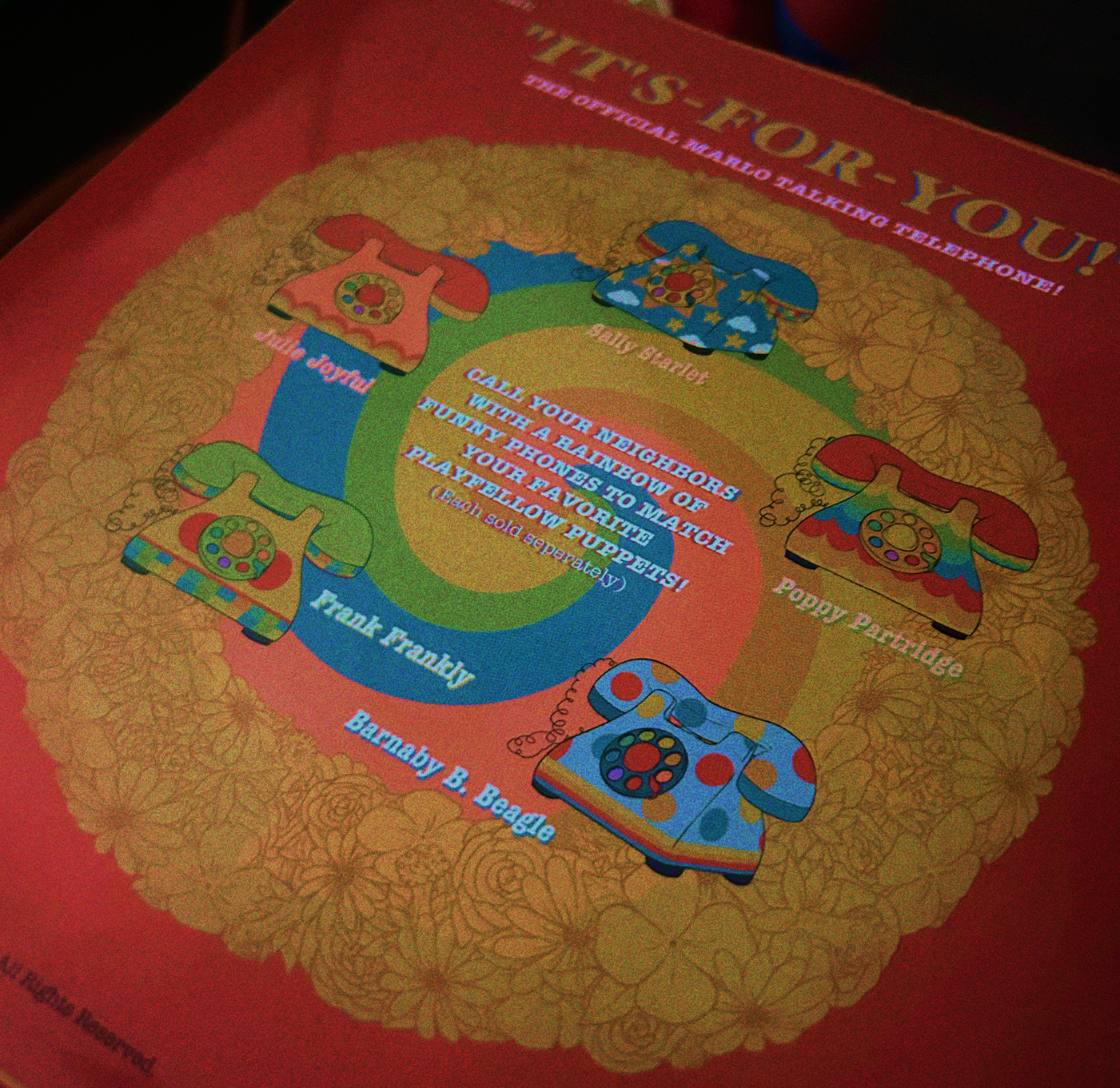 A photograph of the other side of the box, which has an illustration of 5 other phones styled to have elements that evoke Sally, Julie, Poppy, Frank, and Barnaby. Text reads 'Call your neighbors with a rainbow of funny phones to match your favorite playfellow puppets! (each sold seperately)'