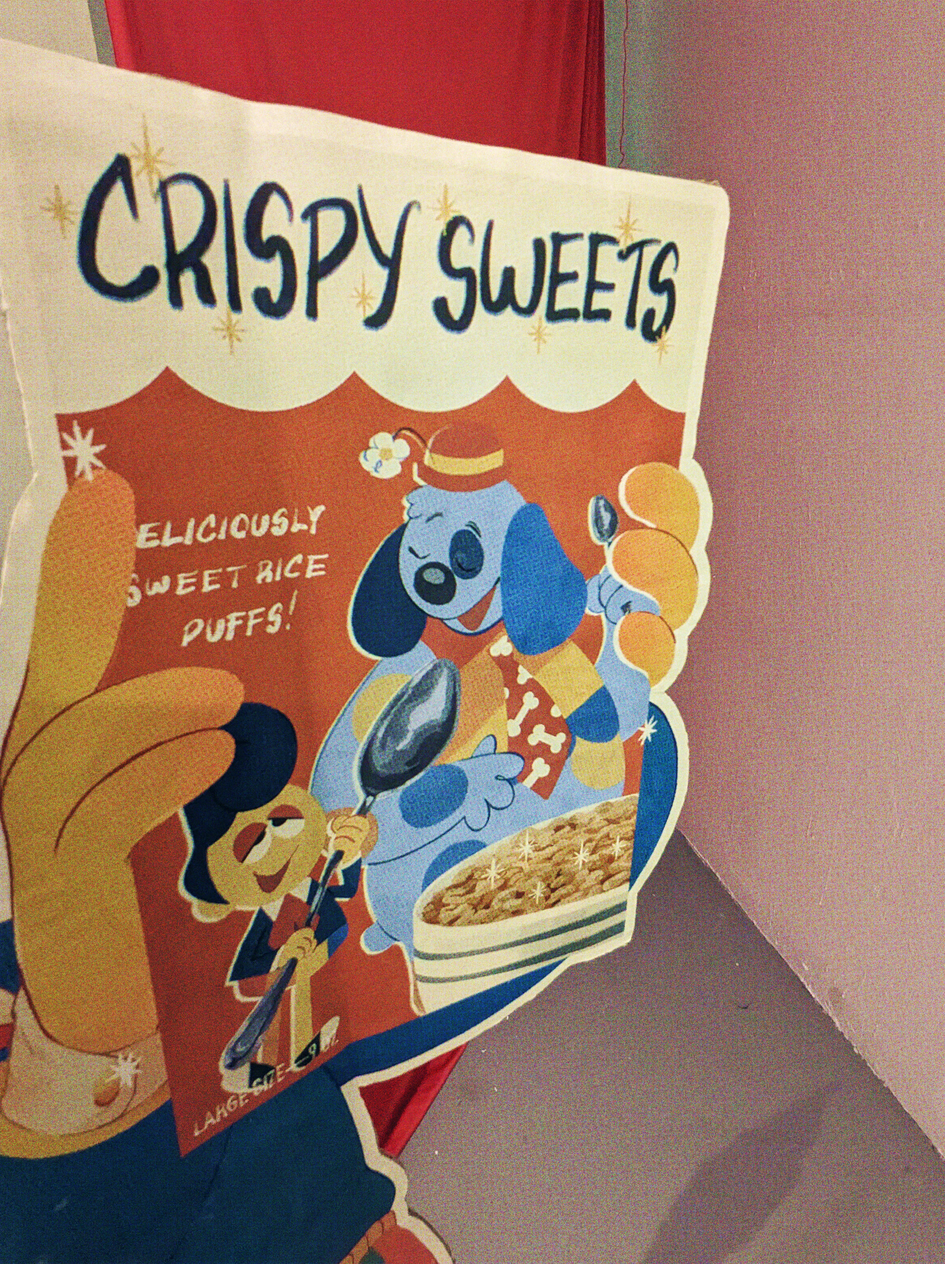 A photograph of a Wally Darling Crispy Sweets advertisement cardboard standee, it's a closeup on the Crispy Sweet cereal he's holding, which has an illustration of he and Barnaby with spoons, and a slogan 'deliciously sweet rice puffs!'
