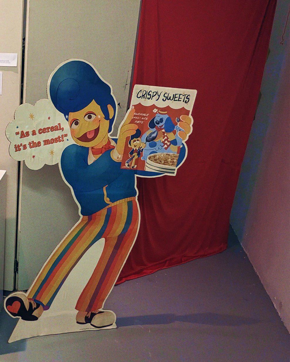 A photograph of a Wally Darling Crispy Sweets advertisement cardboard standee, it's taken from further away, showing that he has written text next to his face that reads 'As a cereal, it's the most!'