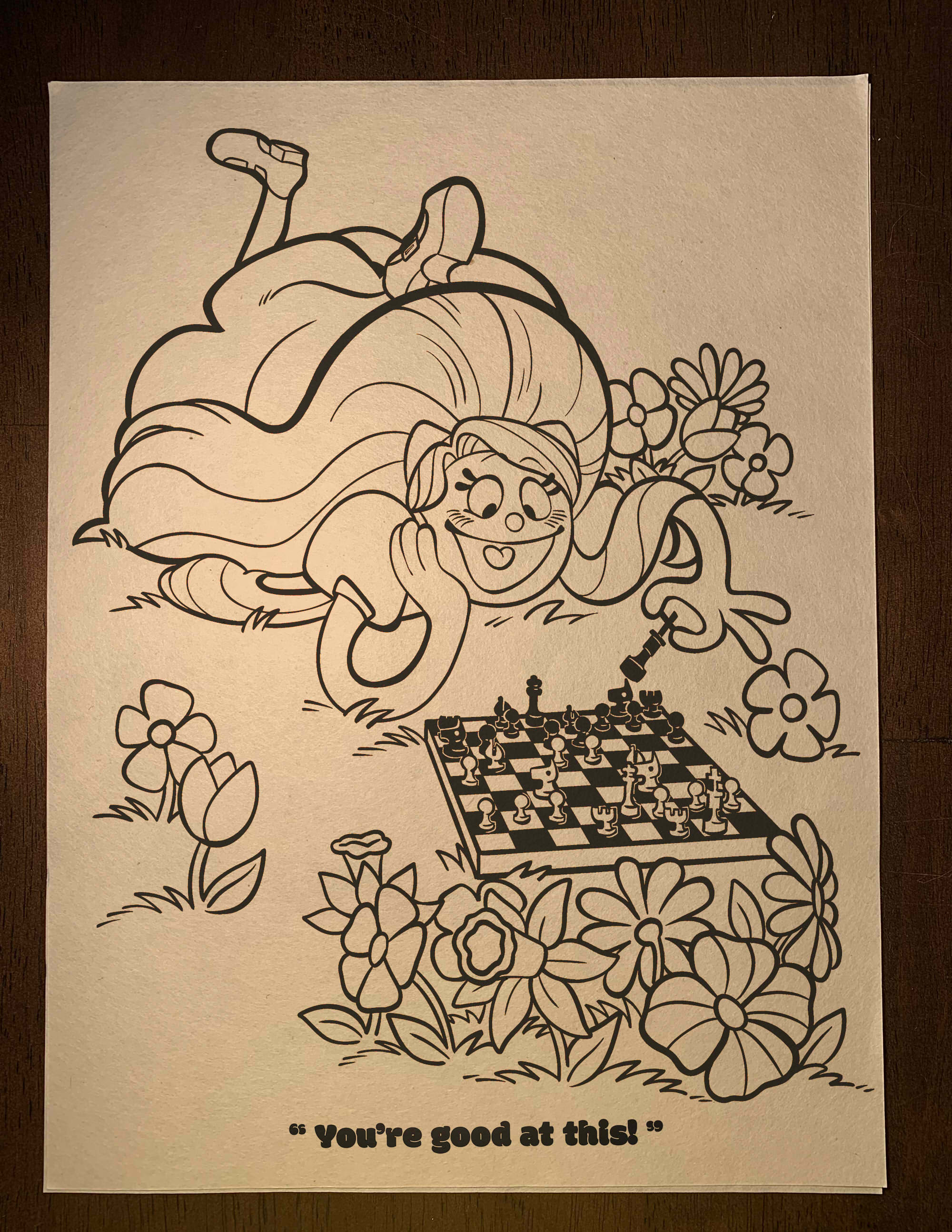 An uncolored page from inside the coloring book showing Julie smiling and kicking her feet as she plays chess against a group of flowers. She says: 'You're good at this!'
