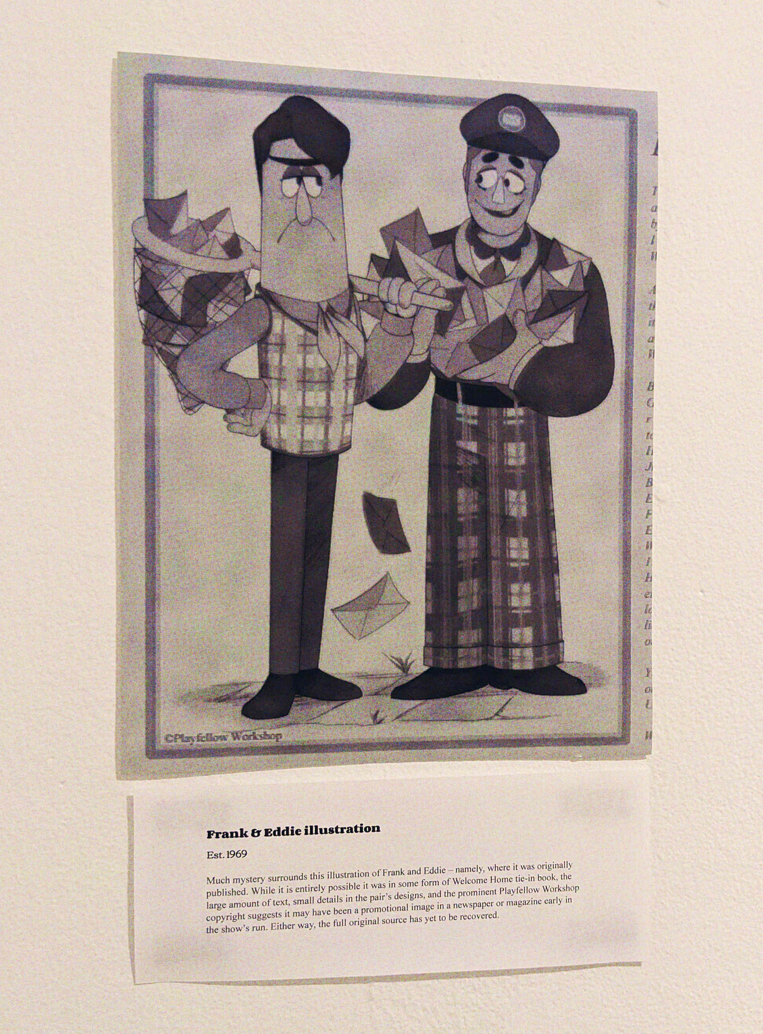 A photo of a wall with an illustration and exhibit card. The card reads: 'Frank & Eddie illustration. Est. 1969. Much mystery surrounds this illustration of Frank and Eddie - namely, where it was originally published. While it is entirely possible it was in some form of Welcome Home tie-in book, the large ammount of text, small details in the pair's designs, and the prominent Playfellow Workshop copyright suggests it may have been a promotional image in a newspaper of magazine early in the show's run. Either way, the full original source has yet to be recovered.' The illustration shows Frank and Eddie standing next to eachother, Frank has a stern expression and is holding a bug net that's filled with envelopes. Eddie meanwhile sheepishly smiles as he holds a ton of envelopes, some fluttering to the ground. There's a Playfellow workshop copyright in the left bottom corner, and cut off to the right is what appears to be a tiny sliver of a look into a wall of text