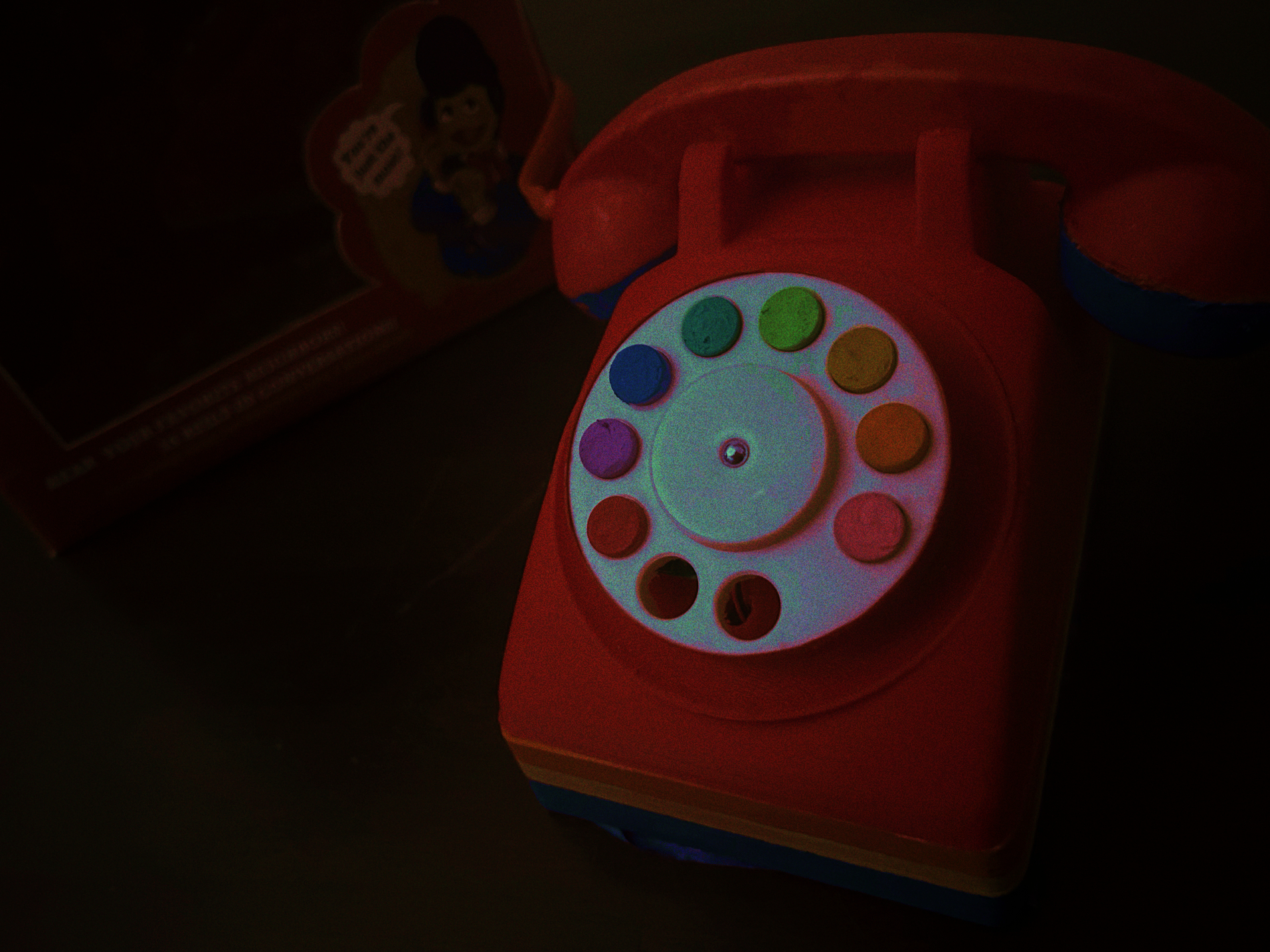 A photograph of a toy telephone, it's red, with yellow and blue accents. There are 8 buttons, of which are a rainbow of colors (pink through purple, back to red).