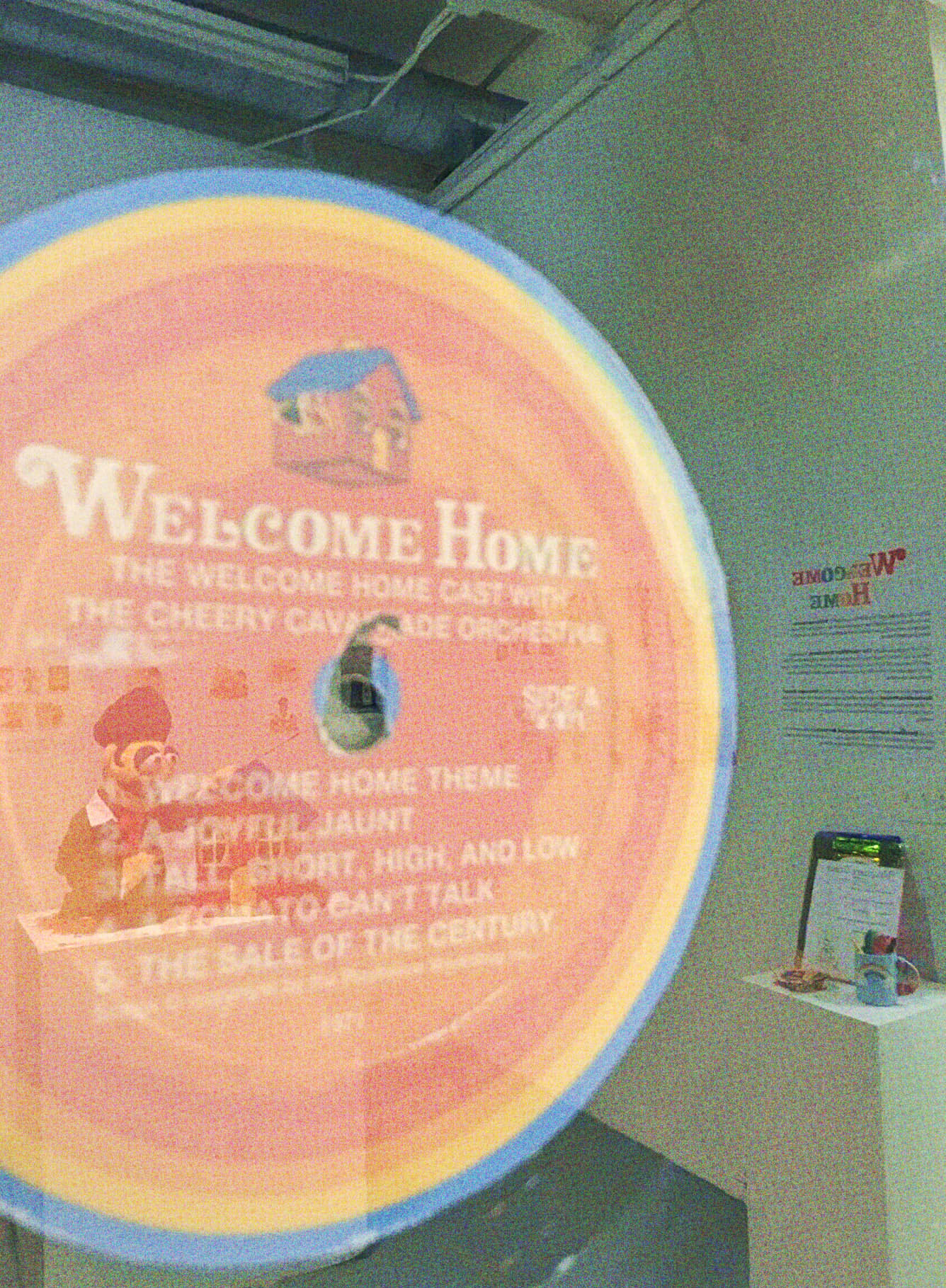 A closeup on a vinyl record's center, showing an illustration of Home alongside the series logo, the further text reads 'The Welcome Home Cast With the Cheery Cava(illegible)ade orchestra Side A. 1. Welcome Home Theme. 2. A Joyful Jaunt. 3. Tall, short, high, and low. 4. A tomato can't talk. 5. The sale of the Century.' The vinyl is framed, with the Wally puppet replica being visible in the glasses reflection, as well as the table of worksheets.