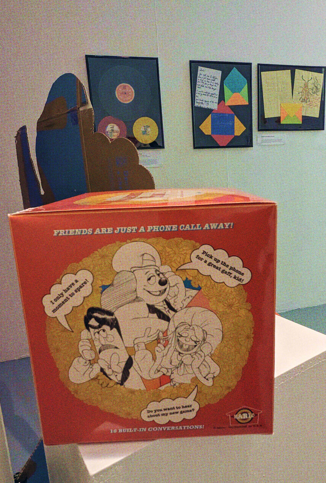 A photograph of the opposite side of the toy telephone's box, showing that the wall behind to it has frames of a vinyl record set, and the early shown Frank and Howdy letters, as well as the Wally crispy sweets cutout. The box however shows the slogan 'Friends are just a phone call away!' With illustrations of Barnaby, Frank, and Julie, all on the phone. Barnaby wears a smug expression and says 'Pick up the phone for a great gaff, kid!' Frank has a exhasperated look and says 'I only have a moment to spare!' while Julie twirls her hair and closes her eyes, smiling, as she says 'Do you want to hear about my new game?' it again touts having 16 built-in conversations, and the Marlo brand logo is visible in the corner.