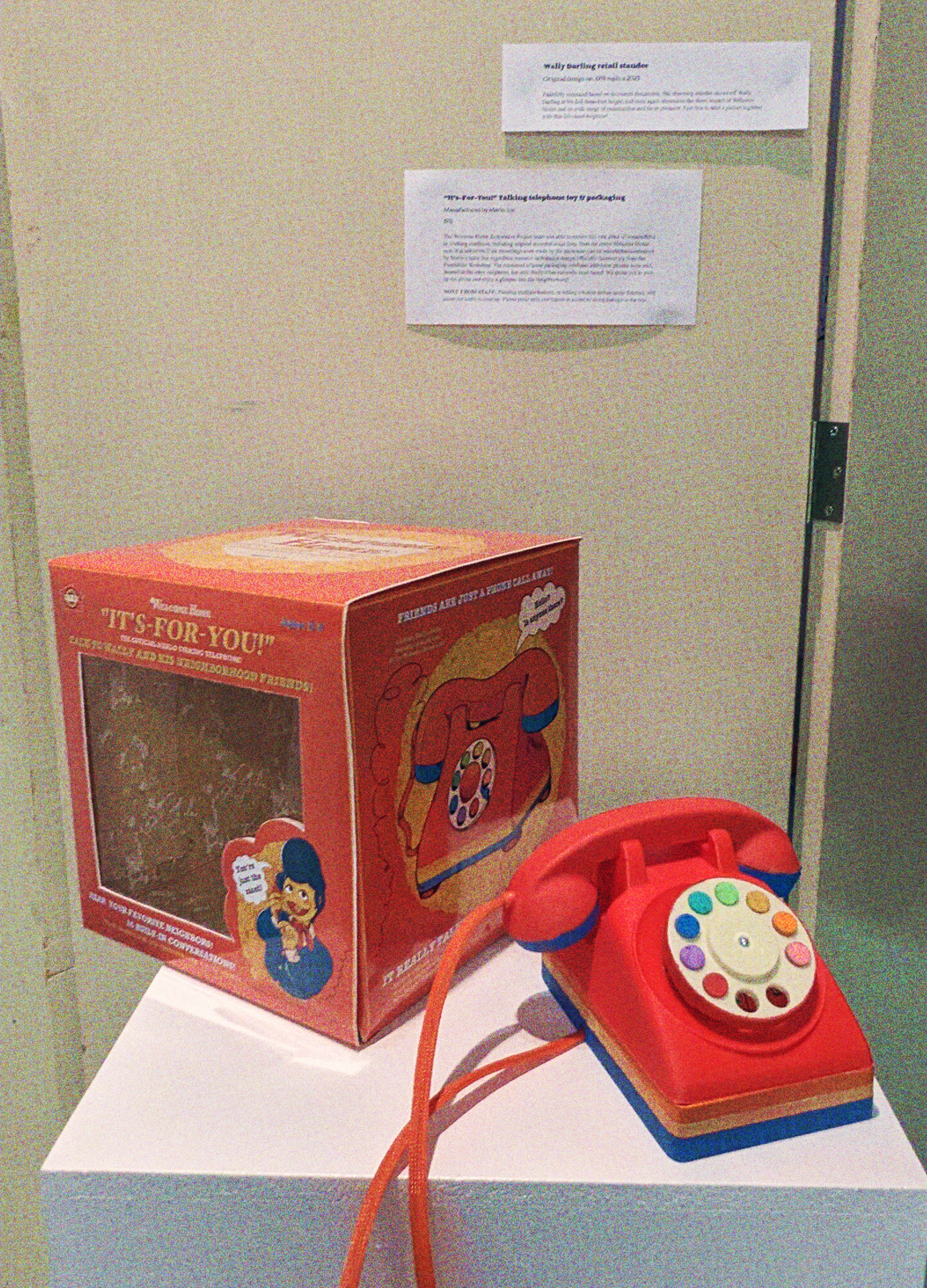 A photograph of the toy telephone and box on display, theres an exhibit card behind it that reads 'It's-For-You!' talking telephone' and another exhibit card for the Wally crispy sweets standee titled 'Wally Darling retail standee' The other text is too blurry to read.