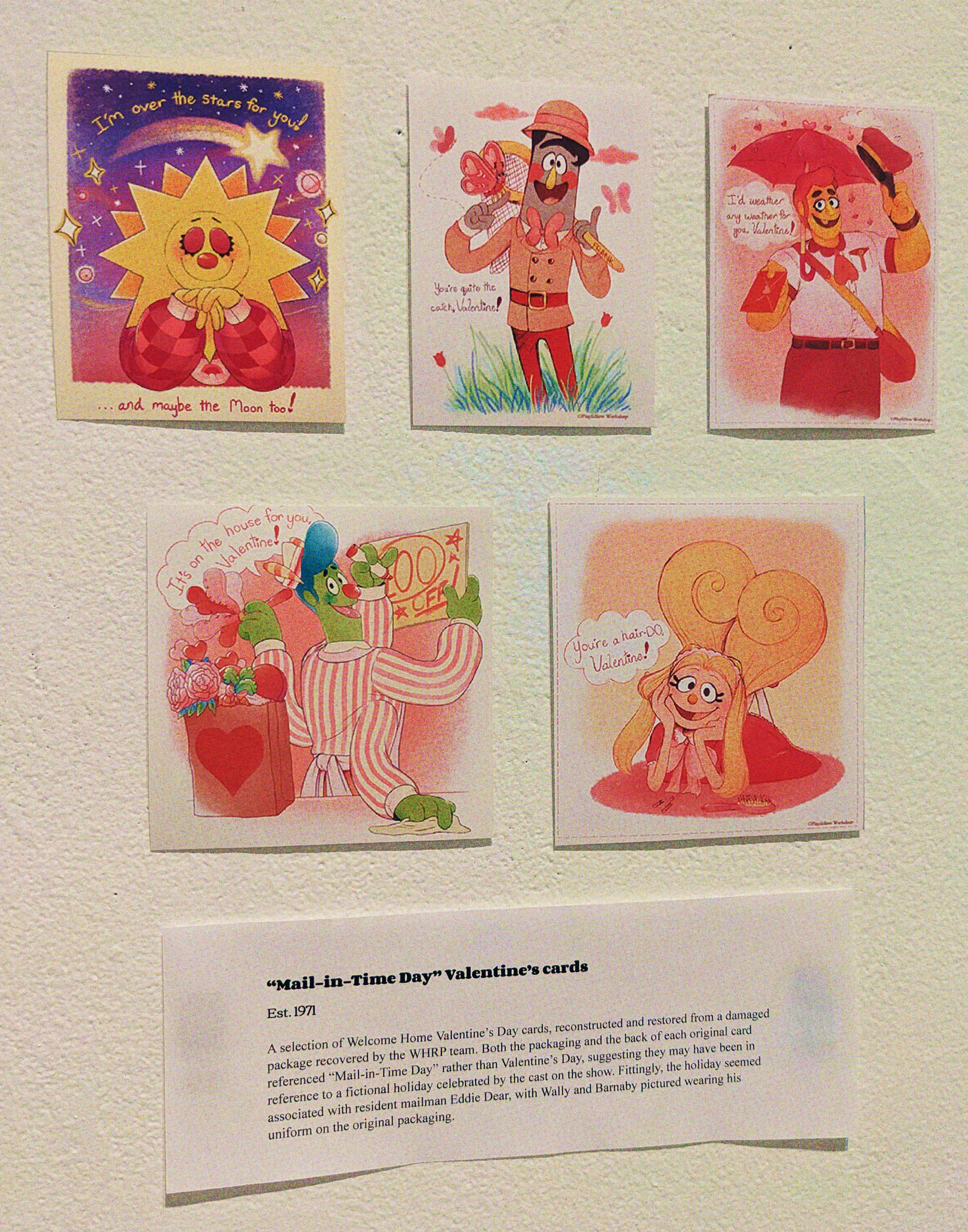A wall displaying 5 valentines, alongside a exhibit card that reads: 'Mail-inTime Day Valentine's cards Est. 1971. A selection of Welcome Home Valentine's Day cards, reconstructed and restored from a damaged package recovered by the WHRP team. Both the packaging and the back of each original card referenced 'Mail-in-Time Day' rather than Valentine's Day, suggesting they may have been in reference to a fictional holiday celebrated by the cast on the show. Fittingly, the holiday seeed associated with resident mailman Eddie Dear, with Wally and Barnaby pictured wearing his uniform on the original packaging.' The illustrations show 1) Sally smiling and closing her eyes as a shooting star flies in the sky of the background, text reads 'I'm over the stars for you! ...and maybe the moon too!' 2) Frank smiling, holding a bug net in one hand, and has a butterfly perched on his finger on the other. text reads 'You're quite the catch, Valentine!' 3) Eddie tipping his hat while he holds an umbrella and an envelope, he's smiling and the raindrops are drawn as hearts text reads: 'i'd weather any weather for you, valentine!' 4) Howdy smiling as his multiple hands, make a sale sign, dust the wall, and wipe the counter. On the counter is a papr bag with flowers and a chocolate container, text reads 'It's on the house for you Valentine!' 5) Julie smiling, her hair is shaped in a heart. text reads: 'You're a hair DO Valentine!'