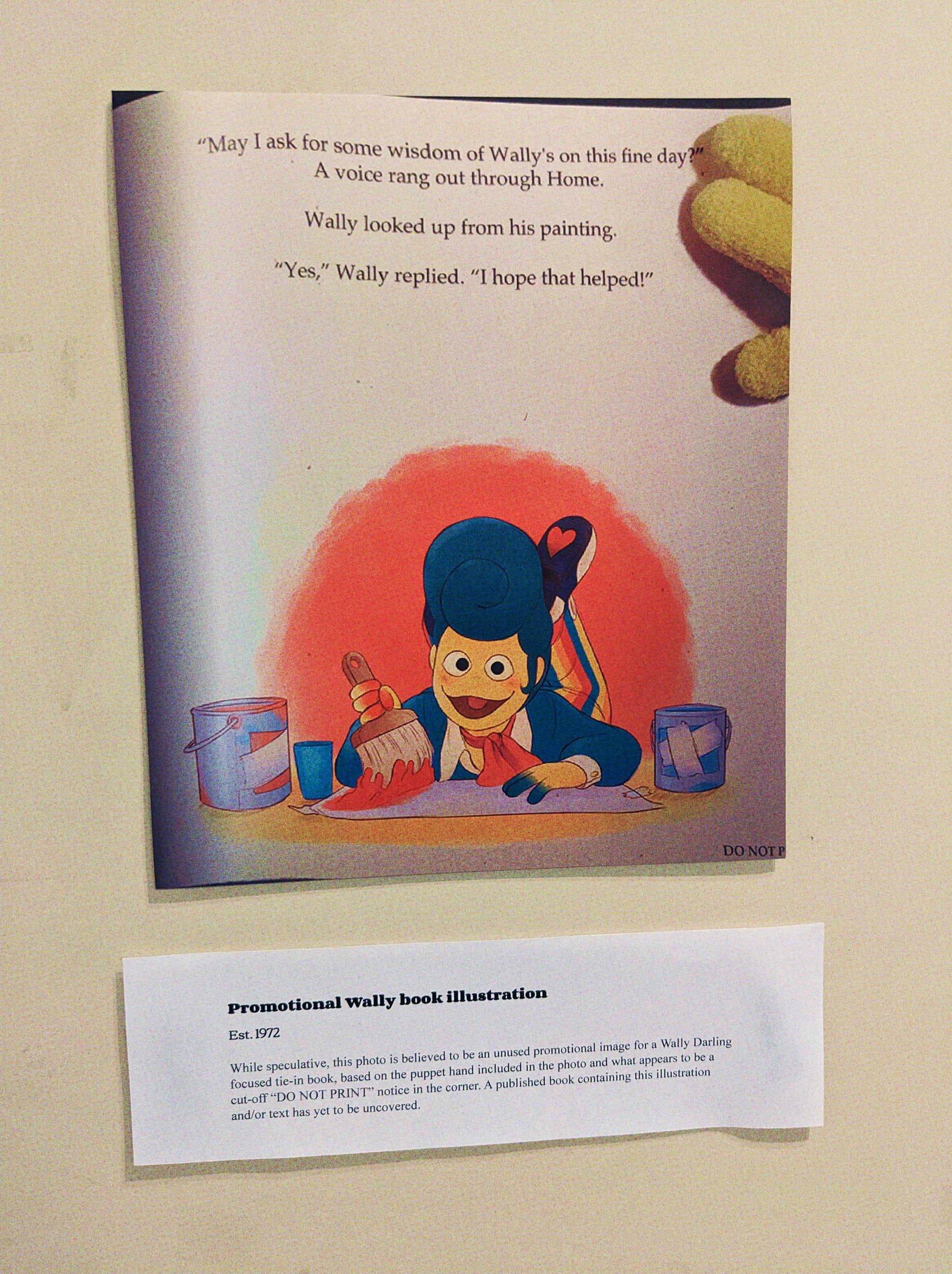 A photo of a wall with a photo and exhibit card. The card reads: 'Promotional Wally book illustration Est. 1972. While speculative, this photo is believed to be an unused promotional image for a Wally Darling focused tie-in book, based on the puppet hand included in the photo and what appears to be a cut-off 'DO NOT PRINT' noticed in the corner. A published book containing this illustration and/or text has yet to be uncovered.' The photo shows an illustration of Wally laying on the ground, painting and smiling. Text reads ''May I ask for some wisdom of Wally's on this fine day?' A voice rang out through Home. Wally looked up from his painting, 'Yes,' Wally replied. 'I hope that helped!' In the top right corner is a glimpse at a Wally Darling puppet hand as if he's the one holding the page open, and in the bottom right corner is a tiny 'DO NOT P-' that gets cut off.