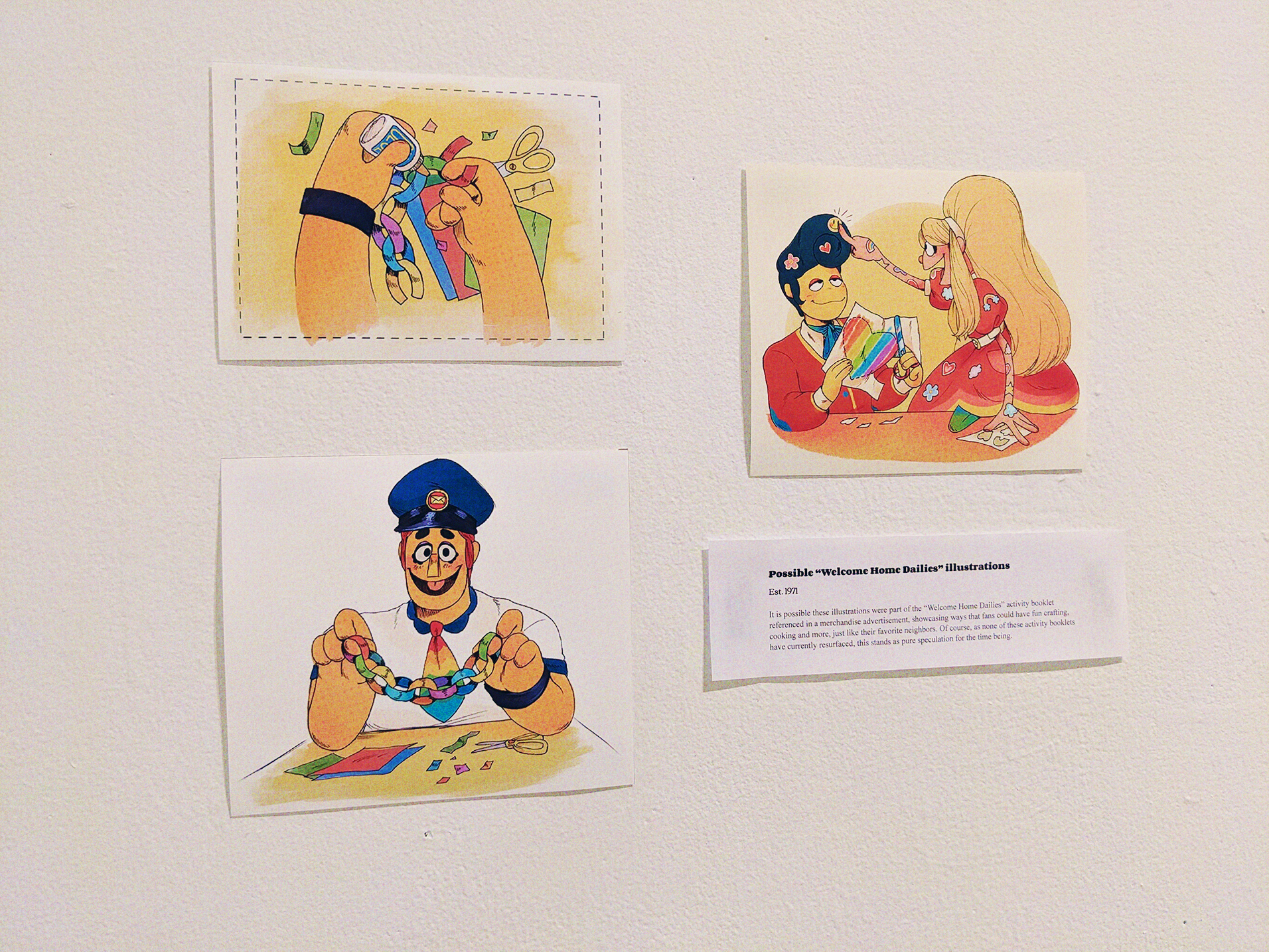 A photo of a wall with three illustrations, and an exhibit card. The card reads: 'Possible 'Welcome Home Dailies' illustrations. Est. 1971. It is possible these illustrations were part of the 'Welcome Home Dailies' activity booklet referenced in a merchandise advertisement, showing ways that fans could have fun crafting, cooking, and more, just like their favorite neighbors. Of course, as none of these activity booklets have currently resurfaced, this stands as pure speculation for the time being.'  The three illustrations show Eddie cutting out and gluing paper into a paper chain garland, and holding it up while smiling. Another shows Julie putting stickers on Wally (while she's already covered in them) while Wally trims a rainbow heart out of a piece of paper.