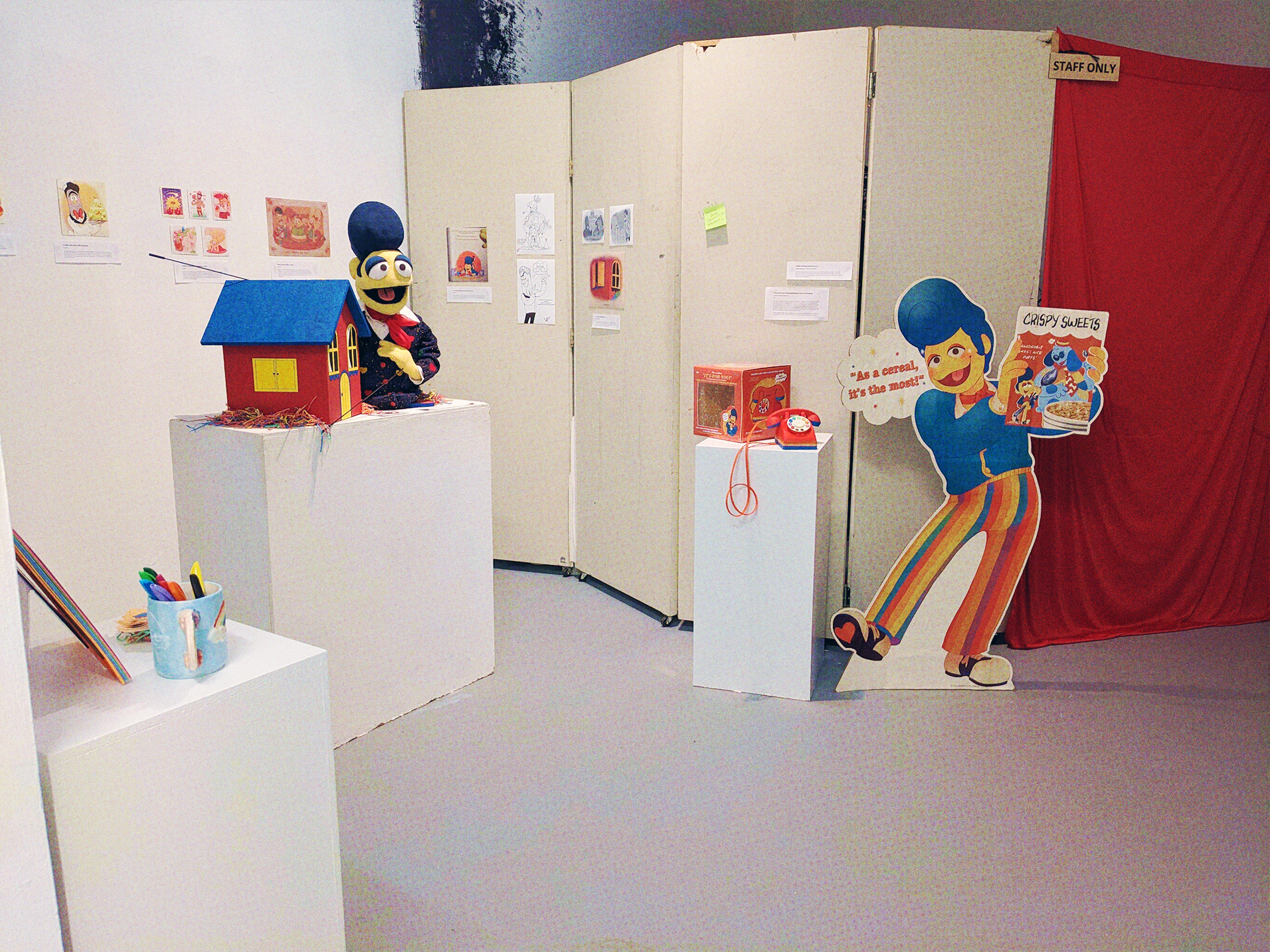 A overview of the exhibit, showing several things taped to the wall, the Wally Standee, the toy telephone, and a Wally puppet next to a large Home. To the right is a red curtain that has a sign reading 'Staff Only'
