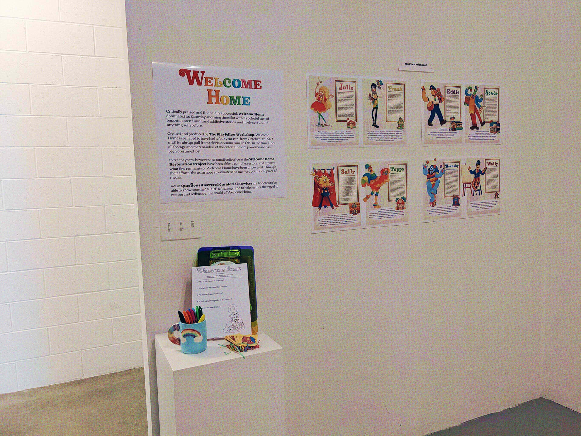A photograph of one of the exhibit walls, the one with pages of all the residents. Next to it is a card that reads 'Welcome Home, Critically praised and financially successful, Welcome Home dominated its Saturday-morning time slot with its colorful cast of puppets, entertaining and addictive stories, and lively sets unlike anything seen before. Created and produced by The Playfellow Workshop, Welcome Home is believed to have had a four year run, from October 11th, 1969 until its abrupt pull from television sometime in 1974. In the time since, all footage and merchandise of the entertainment powerhouse has been presumed lost. In recent years however the small collective at the Welcome Home Restoration Project have been able to compile restore and archive what few remnants of Welcome Home have been uncovered. Through their efforts, the team hopes to awaken the memory of this lost piece of media. We at Questions Answered Curatorial Services are honored to be able to showcase the WHRP's findings, and to help further their goal to restore and rediscover the world of Welcome Home.' under the card, is a table that has worksheets, a cup with markers in it, and business cards.
