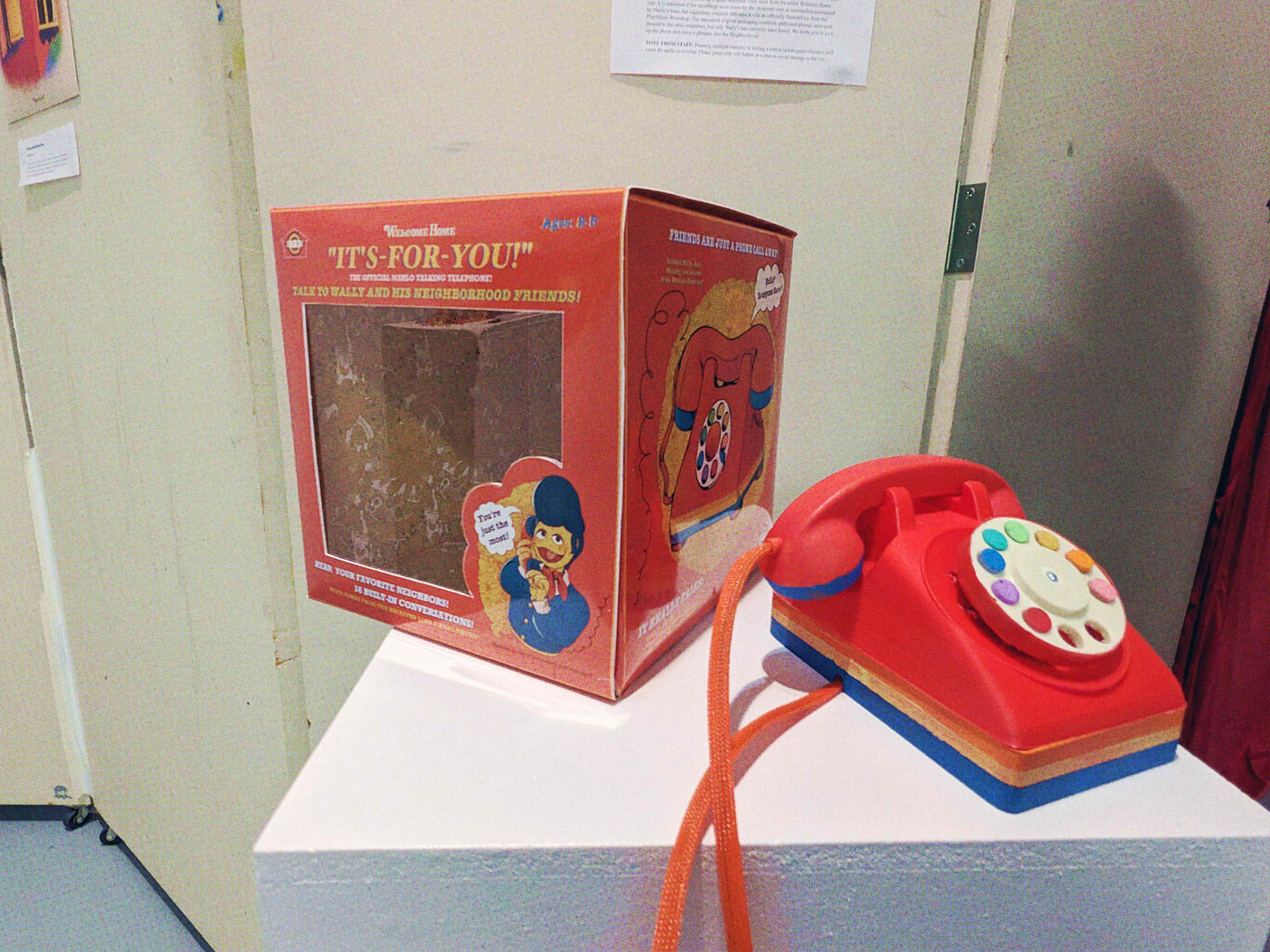 A photograph of a toy telephone alongside it's box. The telephone is red, with yellow and blue accents. There are 8 buttons, of which are a rainbow of colors (pink through purple, back to red). The front of the box reads: 'Welcome Home IT'S-FOR-YOU! The official Marlo Talking Telephone! Talk to Wally and his neighborhood friends! Hear your favorite neighbors! 16 built-in conversations!' Alongside an illustration of Wally on the phone, twirling the cord and smiling, saying 'You're just the most!'