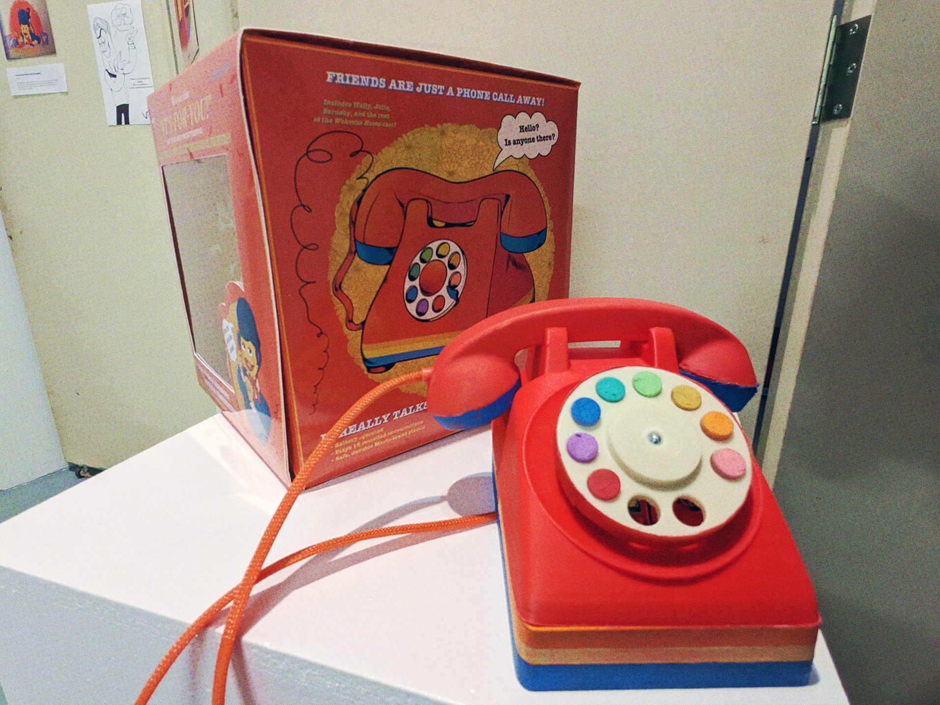 A photograph of the toy telephone alongside it's box, this time showing the phone closer up, and the side of the box. The box has an illustration of the phone, with a speach bubble that reads 'Hello? Is anyone there?' and the slogan 'Friends are just a phone call away!'