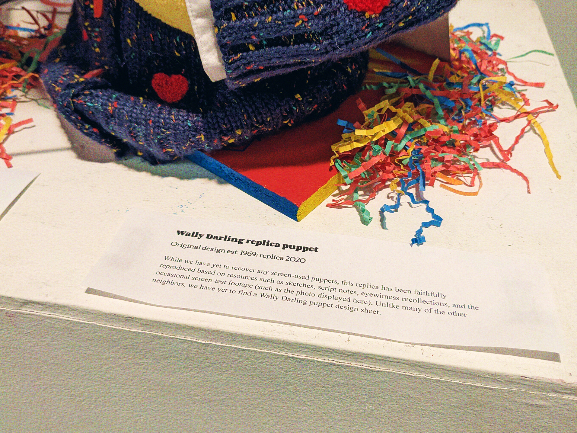 A closeup on the exhibit card for the puppet, reading: 'Wally Darling replica puppet. Original design est. 1969: recplica 2020. While we have yet to recover any screen-used puppets, this replica has been faithfully reproduced based on resources such as sketches, script notes, eyewitness recollections, and the occasional screen-test footage (such as the photo displayed here). Unlike many of the other neighbors, we have yet to find a Wally Darling puppet design sheet.'